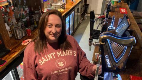 Megan stands behind the bar, smiling up at the camera. Behind her are bottles of alcohol and she stands next to the handles to pull pints. She has shoulder length brown hair and wears a salmon coloured hoodie with white writing on.