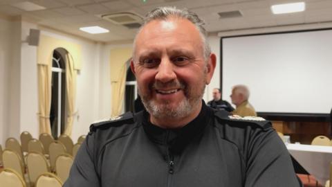 middle aged man with grey hair and a black shirt he is in a room with rows of chairs behind him.