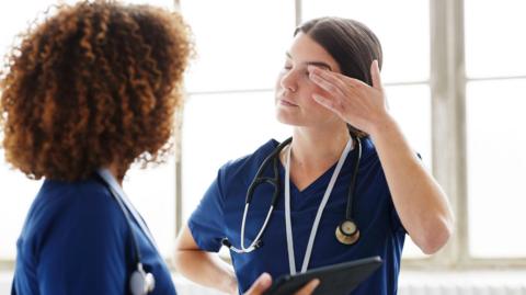 Two women in nurses scrubs. One of them has frilly hair with her back turned, and the other is facing towards the camera, but is touching her eye, looking exhausted. There are windows behind them.