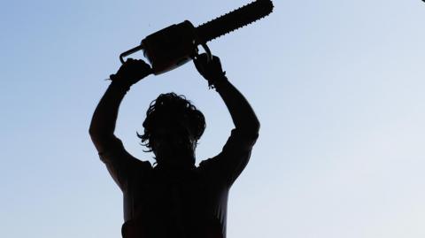 A silhouetted image of somebody dressed as Leatherface holding a chainsaw above their head
