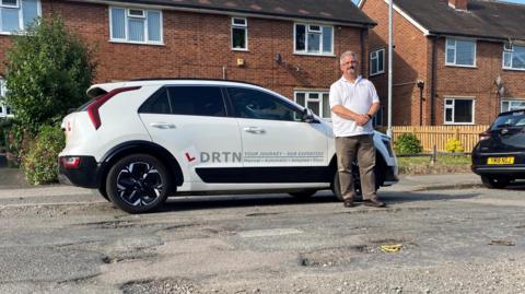 An image of driving instructor Paul Whitburn, standing in front of his car on a road with several potholes.