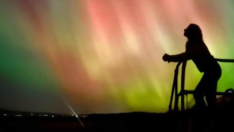 Green, red white and yellow beams of light can be seen across the night sky. Below is the silhouette of a person watching can be