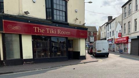 A street view of Rhyl town centre, with The Tiki Room on the left. The yellow building is on a street corner with a red sign running along it with the name 'The Tiki Room'. A doorway and two large windows are seen below with a bay window on the first floor.