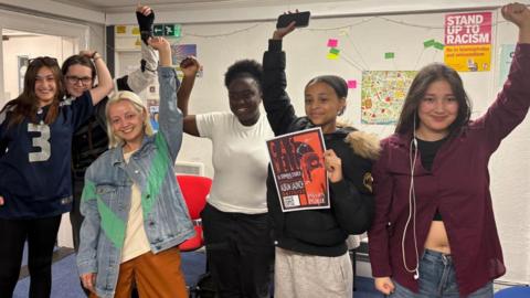 Young women are celebrating the news of their funding with their arms in the air. There are six of them, one is holding a poster, another has headphones hanging out of her ears. They are in a room with a white wall covered in posters. 