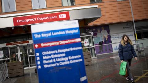 A view of the front entrance to the Royal London Hospital's emergency department on a wet day. The building is clad is long brown tiles, with a protruding steel eave which reads: Emergency Department A&E. A woman carrying a green plastic bag is on the right of the image, and blue hospital signage is out of focus in the image foreground, which reads: The Royal London Hospital.