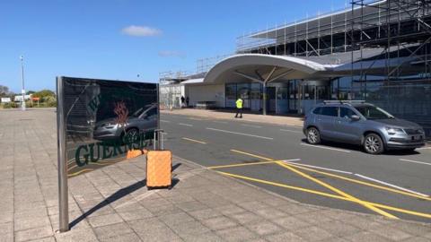 A car drives part a small modern airport. An orange suitcase sits in front of shiny Welcome to Guernsey sign.