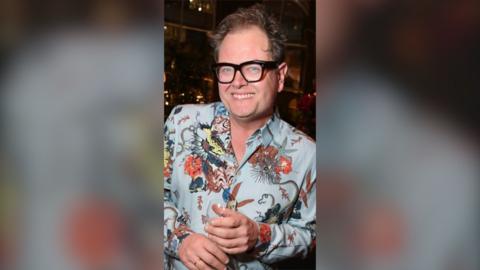 Alan Carr smiles at the camera. He is wearing a pale blue shirt with an elaborate floral pattern on it.