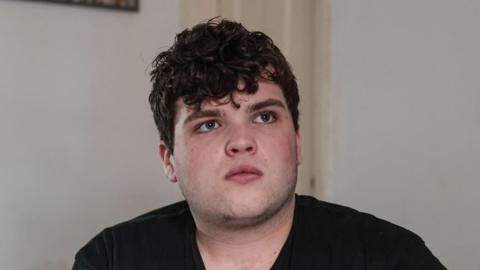 Dylan Cullen poses for picture while sat on a sofa. He wears a black Vans shirt and white walls are behind him.