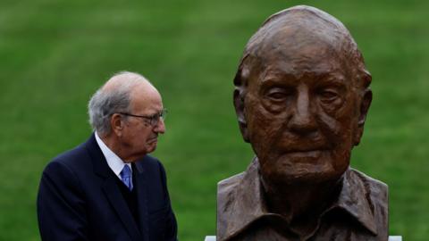 George Mitchell, with short grey hair, wearing glasses, a white shirt, purple tie and navy suit. He is looking to the right at a bust depicting himself.