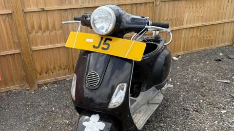 A black scooter is parked on gravel next to a wooden fence. It has the registration plate attached to its handlebars - it reads J5.