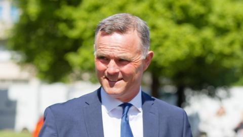 Richard Evan Rhys Williams is pictured leaving Cardiff Crown Court. He is smiling through the sunshine, squinting slightly. He has grey short hair and wears a navy suit, white shirt and navy tie.