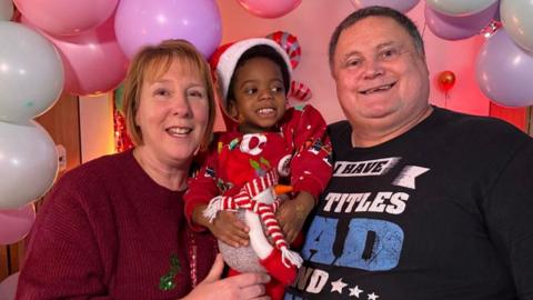A woman with blonde hair is wearing a red top and holding a child dressed in a Santa outfit. On the right is a man with a black top smiling.