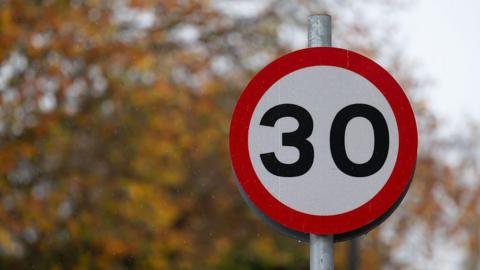 A red and white circular 30mph sign. Autumnal trees can be seen in the background. 