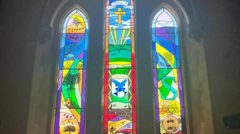 A three-panel stained glass window. It includes drawings of plants, tractors, and a dancer. A cross is featured at the top in the middle panel. There are blue fish, rays of sunshine, and green fields also incorporated into the glass. The window uses a variety of bright colours in abstract patterns.