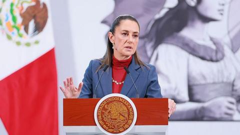 President of Mexico Claudia Sheinbaum speaks during the daily morning briefing at Palacio Nacional in Mexico City, Mexico. She is pictured mid-remarks, wearing a dusty navy blue blazer over a red turtleneck. She is standing behind a wooden podium that features the official seal of Mexico. To her left is the Mexican flag. 