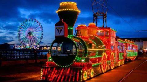 Illuminated heritage tram on Blackpool promenade at night with Central Pier in the background and the Big Wheel lit up.