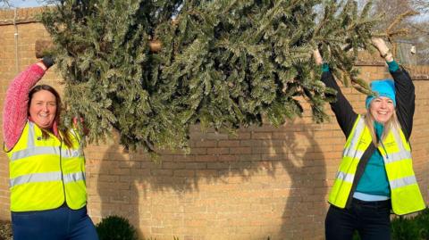 Two women holding a Christmas tree above their heads and smiling. They are standing in front of a brick wall and wearing hi-vis jackets.