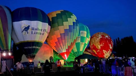 Five multi-coloured hot air balloons that are in a field and preparing to take off as part of a night-time balloon festival. The sky is dark blue in the background. 