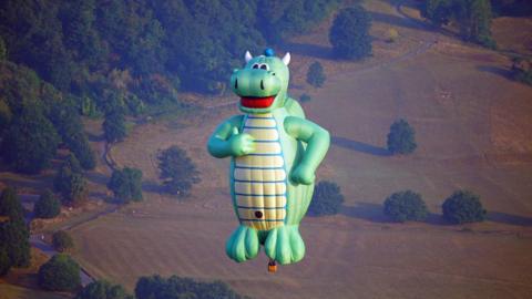A green dragon-shaped hot air balloon with a yellow belly hangs above Bristol in the pale morning sun. The dragon has a large red mouth smile on his face and is holding his thumb up, with his other hand on his hip. Behind him is a field, brown from the summer sun, dotted with mature trees on the edge of woodland.