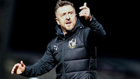 Port Vale boss Jon Brady, wearing a black club gilet, club gives a thumbs up