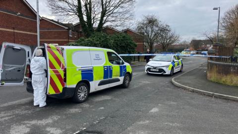 One police car and one police van can be seen on the road, where police tape has blocked off part of the residential street. A person in a full white forensics suit is in the foreground of the picture wearing a mask and head covering. 