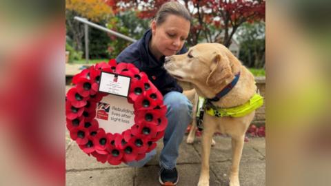 Kelly in a garden, crouching down beside her guide dog Archie who has a harness on and his head is turned towards Kelly. She is holding a large wreath of poppies which says within the circle 'Rebuilding lives after sight loss'