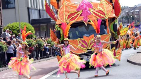 Three female dancers are performing in the middle of a road as part of a parade. They are wearing pink and yellow tutus and carnival clothing, including large feathers on their heads. A bus is behind them covered in orange, pink and gold leaves. The rest of the carnival procession can be seen behind them.