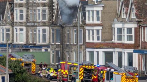 Three fire engines  and other emergency vehicles can be seen in front of a three storey building. There is smoke coming out of the roof.