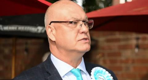 Ian Cooper in a suit and turquoise tie, wearing a rosette, and glasses. Picture taken when he was a candidate. 