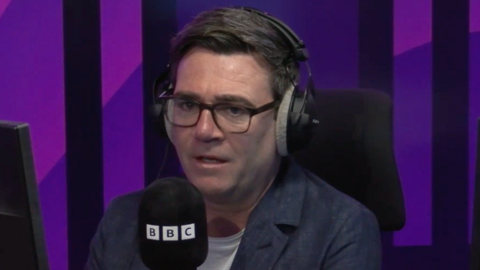 Andy Burnham is interviewed for BBC Radio Manchester. He is wearing headphones and is next to a branded BBC microphone. Burnham wears a white t-shirt, blue jacket and dark-framed glasses.