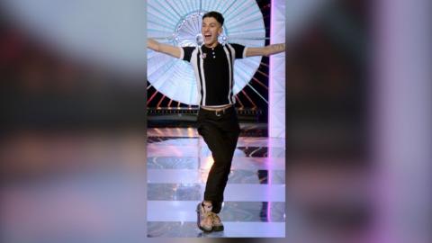 Drag queen Silllexa Diction singing and dancing on the RuPaul's Drag Race UK stage, wearing a black and white polo shirt, with arms outstretched