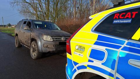 A four-by-four Volkswagen vehicle covered in mud parked on the side of the road with a police car in front of it.