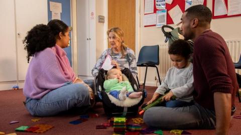 A group of adults with their children having a meeting with a childcare worker. They are in a community centre surrounded by toys. 