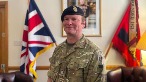 Major David Watson pictured in military uniform, complete with beret. He is in the army barracks and there are flags visible in the background. He is smiling.