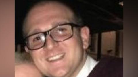 Gary Carson, who is bald, wears glasses and a white shirt. He is photographed smiling into the camera.
