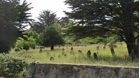 A cemetery with dozens of gravestones. Grass surrounding them has not been cut and is overgrown