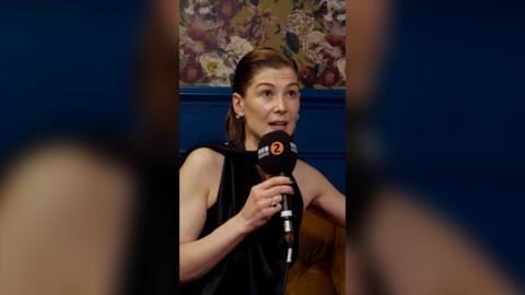 Rosamund Pike wears a black sleeveless dress, holding a Radio 2 microphone.