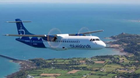 A white and blue, Blue Islands aircraft flying over the Jersey countryside with the ocean in the background. 