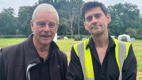 Mike Pinner is smiling at the camera and wearing a pair of glasses, dark polo shirt and fleece jacket. He is standing next to Giles Emery, who is wearing a high vis jacket and black shirt, which is unbuttoned at the neck. They are standing in a field, with tents, cars and trees in the background.