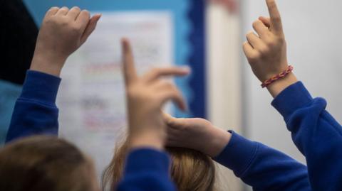 A close-up showing backs of heads of anonymous children wearing blue school uniform jumpers who are holding their hands up in a classroom.