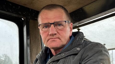 A farmer, a man with short black hair wearing glasses and a coat, sitting in the cab of a tractor on a rainy overcast day.