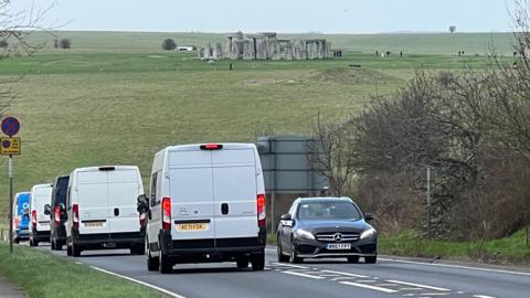 Vans and a car driving along the A303. The Stonehenge monument can be seen in the background on a grey winter's day. Various trees without leaves can be seen to the right of the picture with Stonehenge visitors just visible next to the monument.