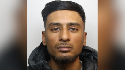 A police custody image of Ali Abbas. He has short black hair, short stubble and wears a black puffer jacket. 