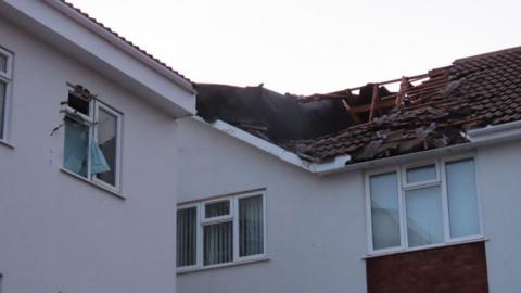 A view of the roof of the building showing part of it missing. The window on the left is also broken. The building is white. The sky is cloudy. 
