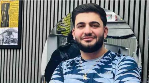 Man Shahram, blue t shirt black hair and beard. Wears watch and necklace and stands in grey painted barber shop.