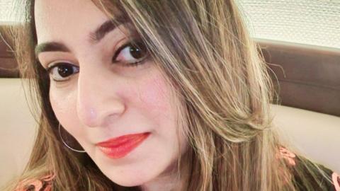 Yaditi Kava has long blonde hair and red lipstick. She is wearing a black and pink dress.