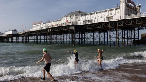 Three people in swimming gear walking into the sea with Brighton pier behind them.