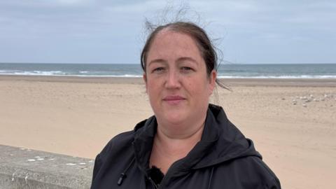 Becca Ringwood is wearing a black coat and standing in front of an empty, flat sandy beach with the sea on the horizon. She has dark hair swept back, strands of which are blowing in the breeze.
