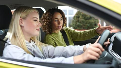 A young woman sitting behind the wheel of a car about to take her driving test. A female driving test examiner is speaking to her first.