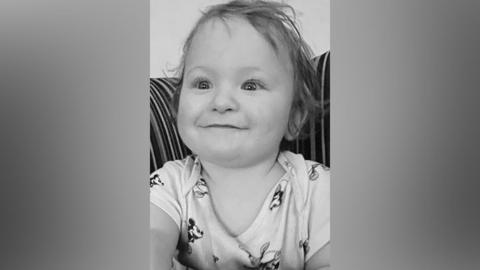 A black and white picture of a smiling Louie Hughes. The baby is wearing a Mickey Mouse vest.